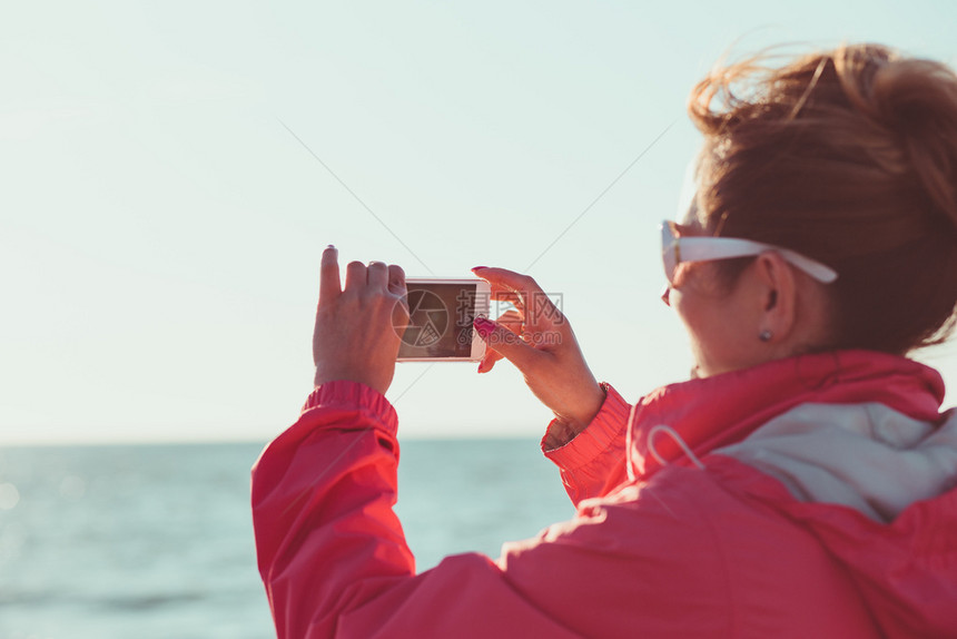 移动的技术年轻女用电话拍照看屏幕站在户外她被阳光照亮背景有浅天和海平面的阳光照耀假期图片