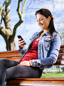 一位美丽的年轻女子坐在长椅上的公园里使用智能手机的照片自然短信春天图片