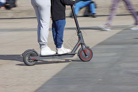 电动滑板车轮母亲和儿子在市广场乘坐电动脚踏车运模糊城市的图片