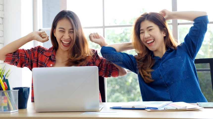 互联网两名年轻女使用笔记本电脑和手臂微笑地脸在家庭办公桌工作成功商业概念在家工作两个年轻的青女使用笔记本电脑和双臂在职的赢图片