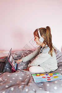 小女孩学龄前幼童习网上解谜游戏玩教育单盘在大流行病检疫期间在家里使用平板电脑和耳机发出声音孩子移动的坐着背景