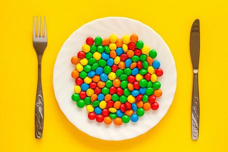饮食多色的糖拖拉机在白色盘子上用刀和叉在黄色背景上好吃图片
