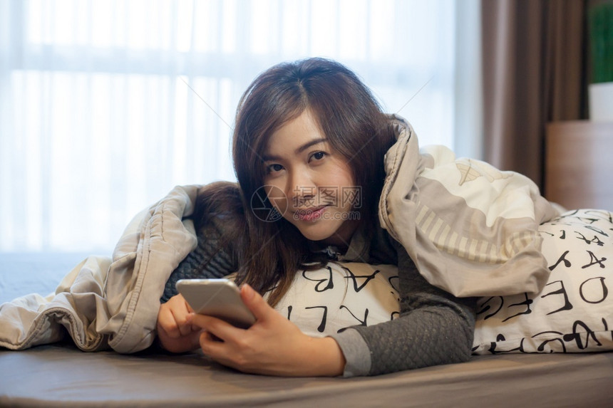 在家里床上用智能手机躺在床上的快乐亚洲年轻女子卧室保持美丽图片