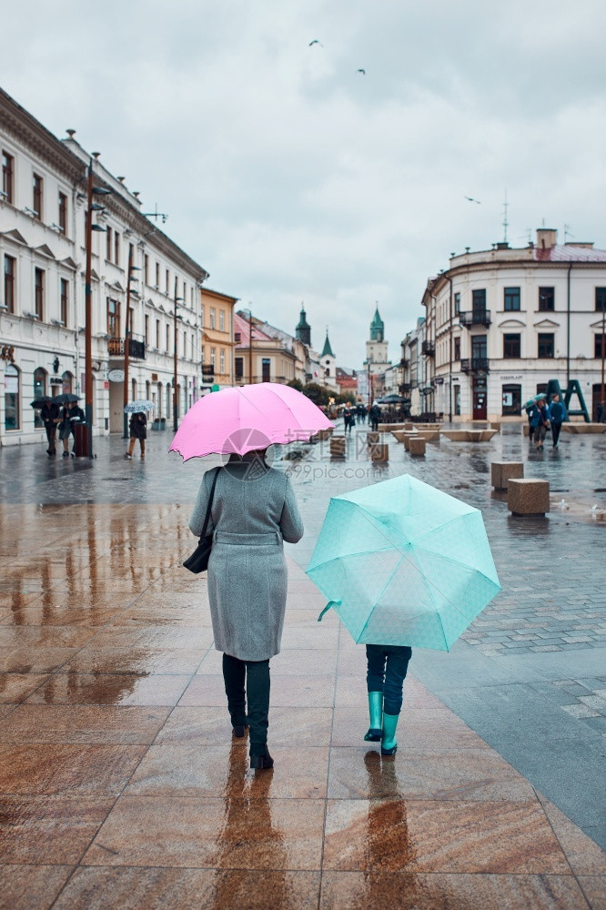 粉色的父母孩子带着粉红和蓝雨伞的母女背视镜在秋雨阴凉的天在市区中行走女人和小孩拿着雨伞在秋的天市区中行走图片