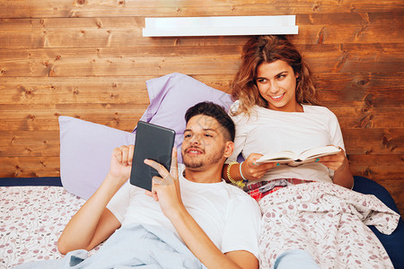 年轻笑着的异恋夫妇躺在床上看书用平板电脑写字床单一种两个都图片