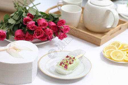 粉色杯子蛋糕摩丝盘子上的冰蛋糕茶壶和托盘上的杯子柠檬片心形盒子和桌上的红玫瑰花束甜桌子背景
