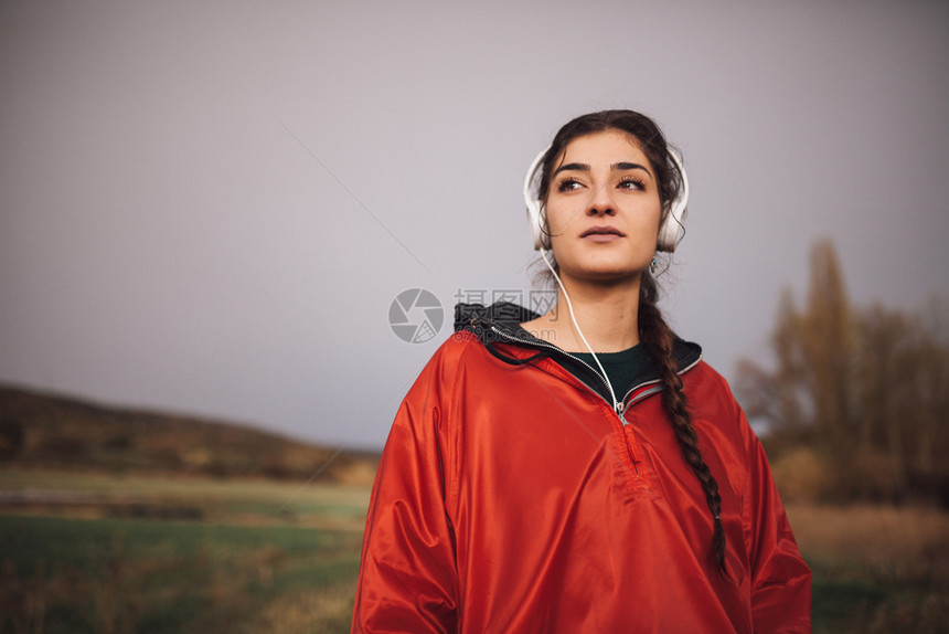 一种场地天气在雨带着衣站的年轻女子在田野上听音乐耳机和图片