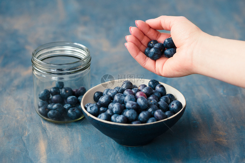 女人把刚从罐子里收集到的蓝莓放一个小碗桌里在背景中漆成蓝色的桌子上新鲜甜陶器图片