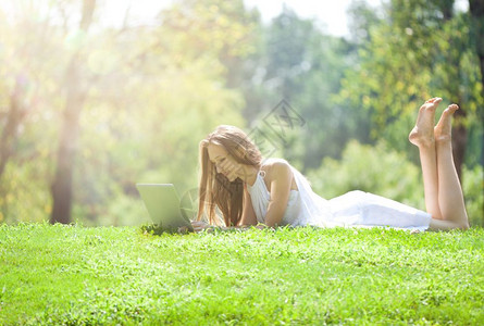 美丽的金发女在明亮的阳光日与笔记本机躺在美丽的绿草地上年轻高加索女士图片