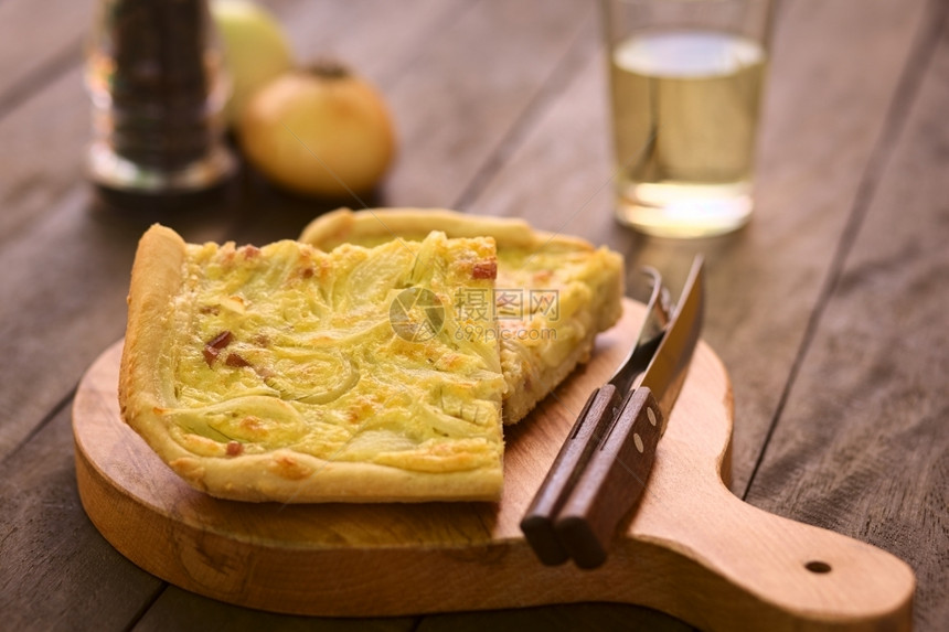 德国传统Zwiebelkuchen或洋葱蛋糕的一块由酵卷面团制成上加洋葱和培根夹在奶油酱中配有木板上的餐具后面是白葡萄酒选择焦点图片