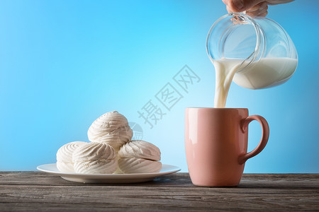 牛奶从一个罐子倒进蓝色背景的杯子健康天然食物的概念早餐牛奶从一个罐子倒进杯寒冷的盘棉花糖背景图片
