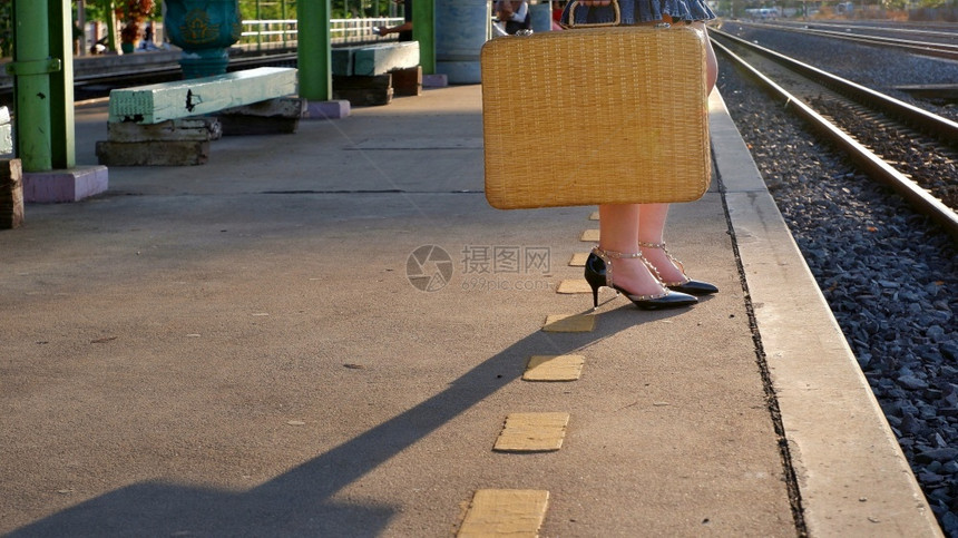 低层年轻女旅行者持有旧式竹手提箱晚上在火车站的平台上等待有阳光和影子的火车厢等候晚间时尚独自的优质图片