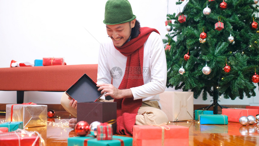 新的年轻亚洲男子在圣诞节庆典晚宴上开圣诞礼物箱时带着快乐的笑容微请享用幸福图片