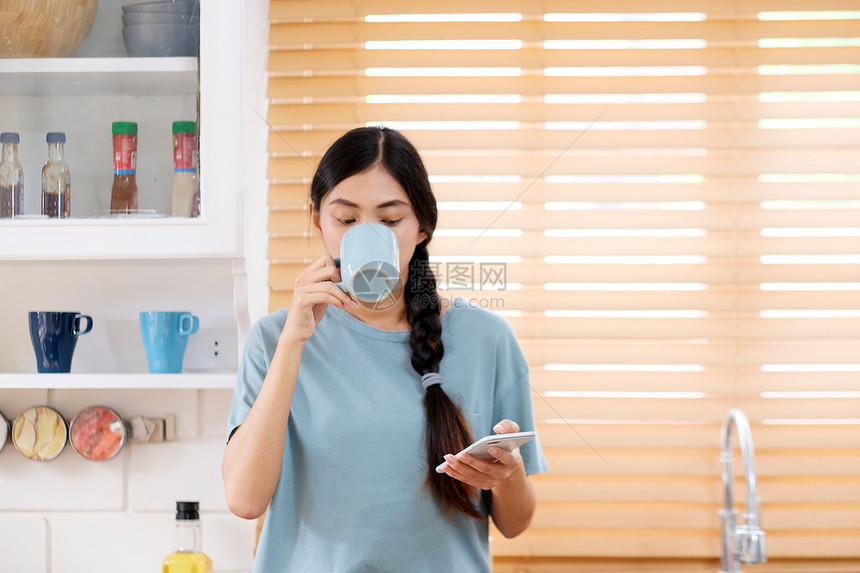 年轻亚洲女使用智能电话同时在厨房背景人和技术生活方式中喝咖啡的烹饪背景生活方式人们尽管互联网图片