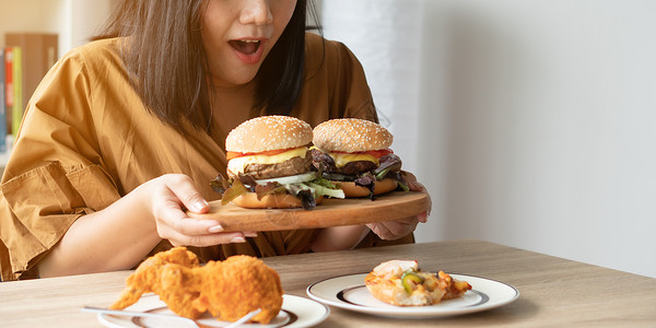 端着火鸡的女孩芝士汉堡饥饿的超重妇女在木板上拿着汉堡包炸鸡和餐桌上边子的披萨狂食饮紊乱概念BED年轻的肥胖背景