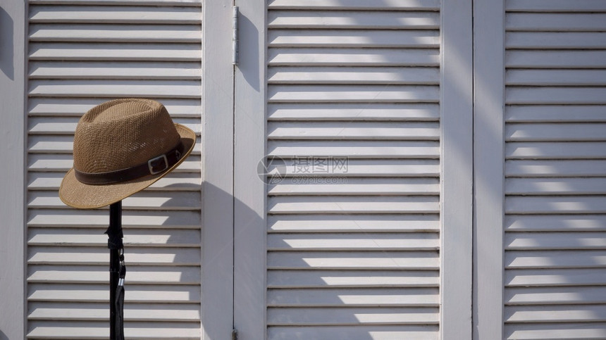 装饰风格控制板软呢帽Fedora帽子表面的阳光和阴影用旧音风格的白色木折叠墙背景上的金属步行棍图片