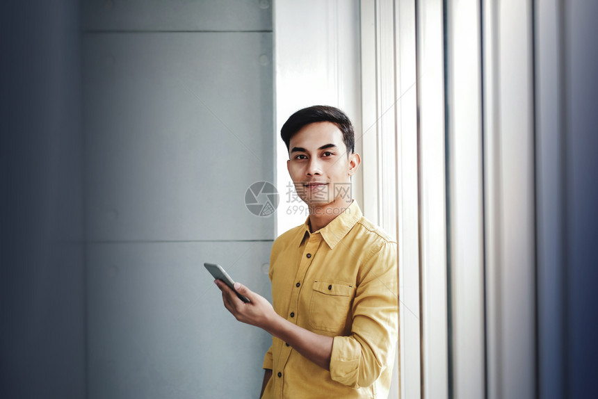 英俊的快乐商人站在办公室窗边用智能手机微笑着看相的肖像男人职业图片