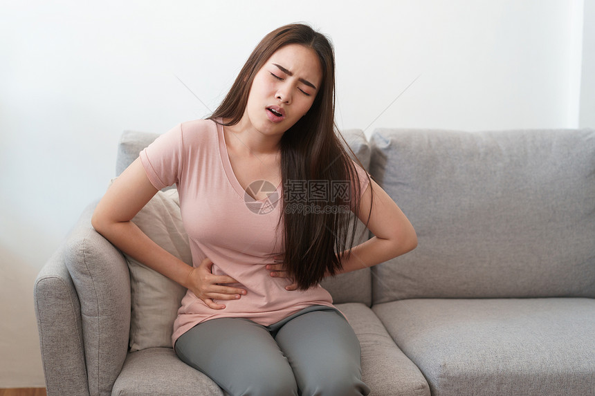 房间健康月经期和疼痛的亚洲女在家中坐沙发上痛苦地坐在沙发上令年轻女抽筋坐着图片