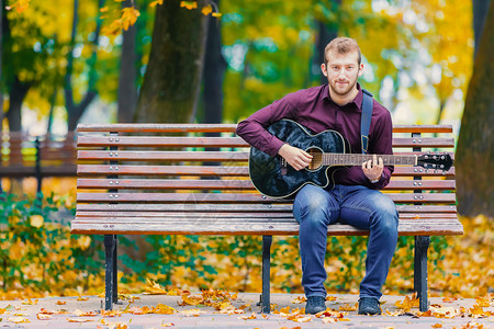 声学坐在长椅上和城市公园弹声吉他的年轻人娱乐玩图片
