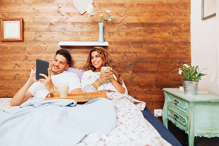 床单人们说谎微笑着的年轻异恋夫妇躺在床上吃早餐用平板电脑一起躺在床上图片