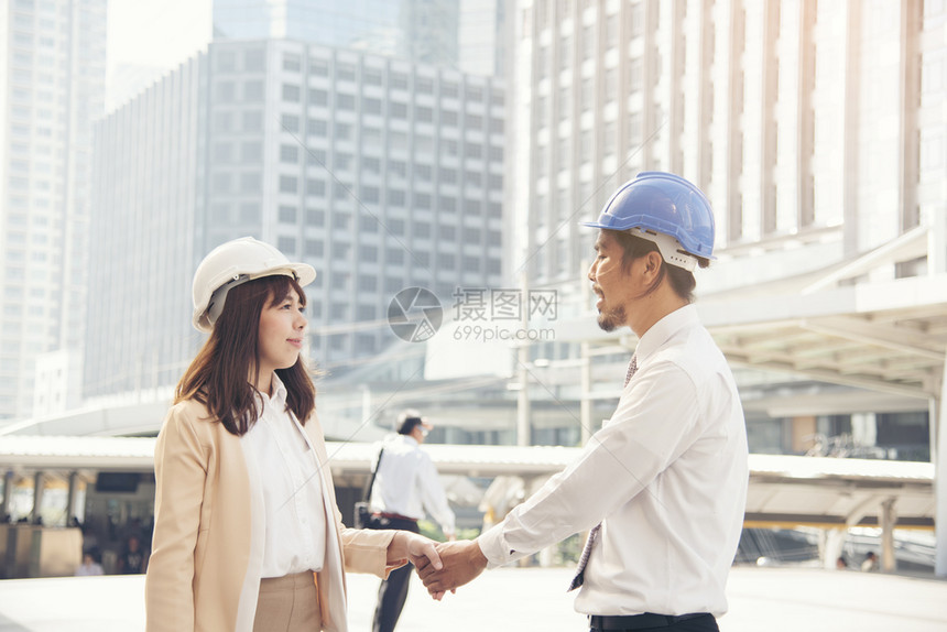 聪明美丽的工程师与英俊头背景城市握手和商业头盔女士图片
