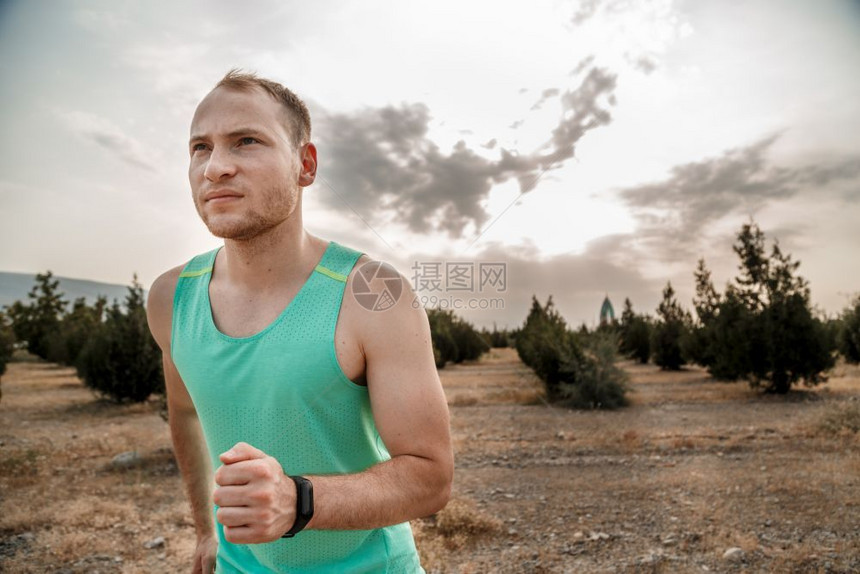 运动的白色穿着蓝T恤和黑短裤的白人男子日落时穿梭粗野地形训练慢跑者图片