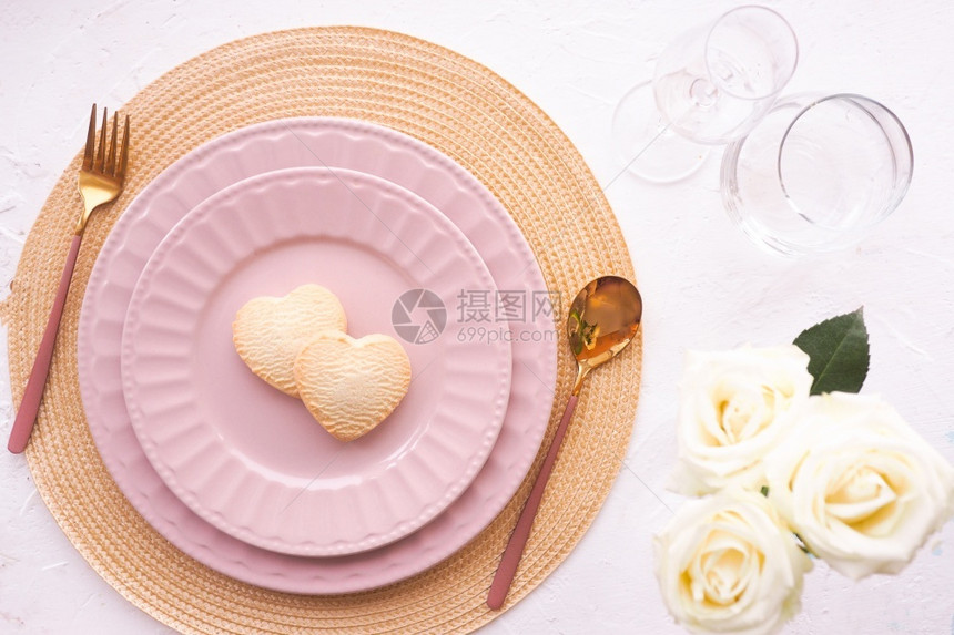 粉色的晚餐桌子上设置巾纸两张粉红板饼干心金礼品盒三个白玫瑰花芽勺子和叉放在一张白色桌上复制平板空间布局花朵图片