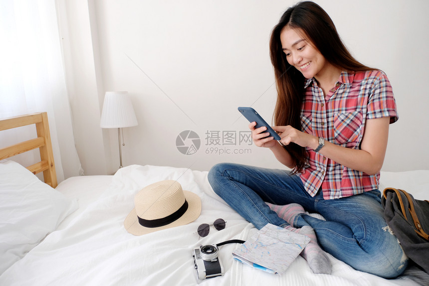 保持背包年轻女旅行者带着和智能手机坐在床上面带微笑的脸模板旅行暑假概念的床边手持和智能机电话图片