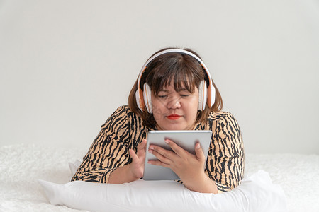 超重女人躺在床上玩平板电脑戴耳机舒适的白天和休息概念轻松声音的弛图片