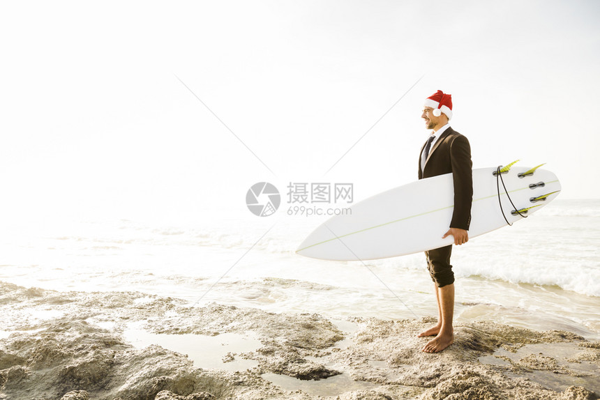 海滩套装爱好身戴圣诞老人帽子并持有冲浪板的商业家图片