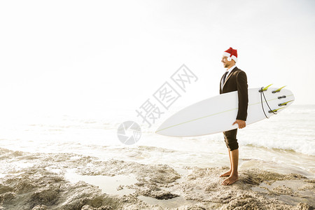 海滩套装爱好身戴圣诞老人帽子并持有冲浪板的商业家图片