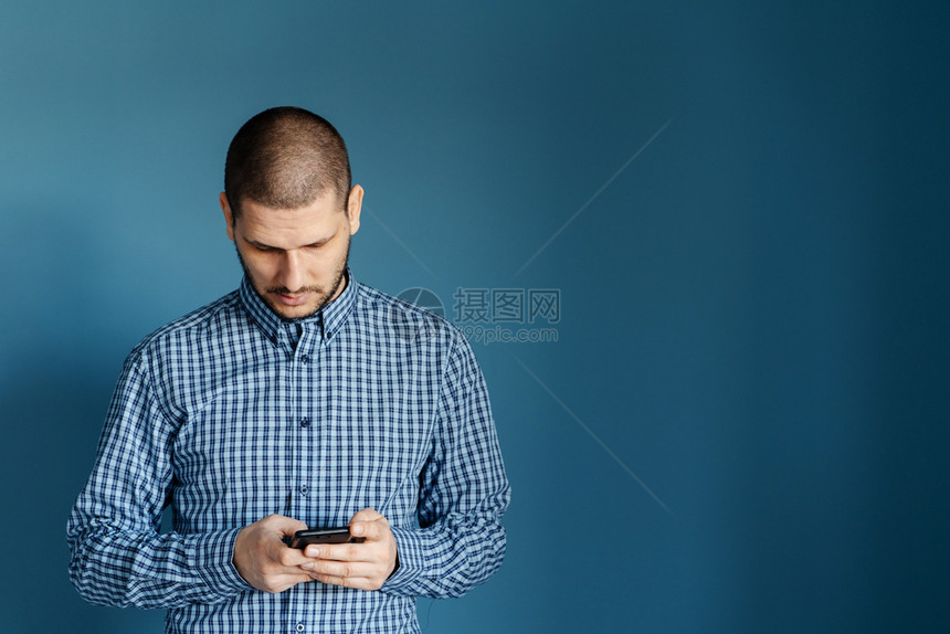 移动的消息传递白种男人穿着衬衫站在蓝背景墙前使用智能手机发送短信或浏览互联网前视图以发送信息短讯或浏览网络前视图在蓝背景墙前面穿图片