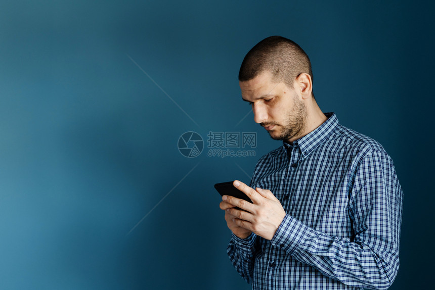 消息自信的蓝色白种男人穿着衬衫站在蓝背景墙前使用智能手机发送短信或浏览互联网前视图以发送信息短讯或浏览网络前视图在蓝背景墙前面穿图片