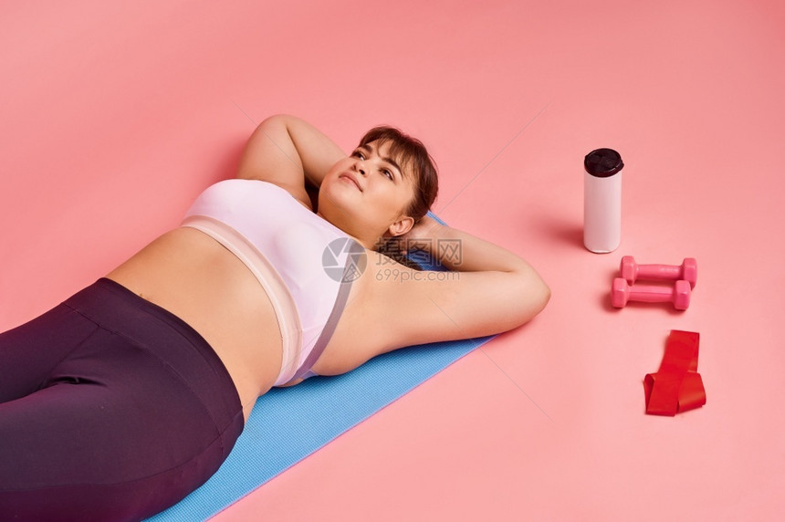 女士超重妇躺在健身床垫上体呈阳粉红色背景肥胖症斗争努力追求健康的生活方式超重妇女躺在床垫上身体呈阳训练年轻的图片