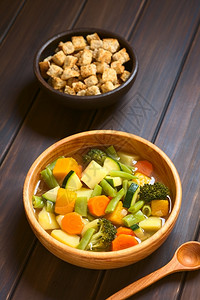 胡萝卜块由青绿豆胡萝卜椰菜花土豆和南瓜制成的木碗蔬菜汤背面有一小碗包条在黑木上用天然灯光拍攝自面包丁健康背景
