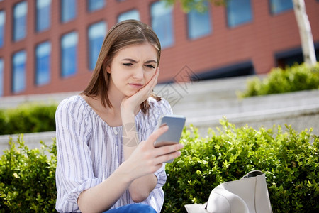 在校园花坛旁坐着玩手机的不开心女孩高清图片