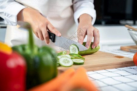 生的亚洲家庭主妇穿围裙用刀在切削纸板上黄瓜和番茄同时准备配料和烹饪供健康餐的午时在家厨房吃切碎美食背景