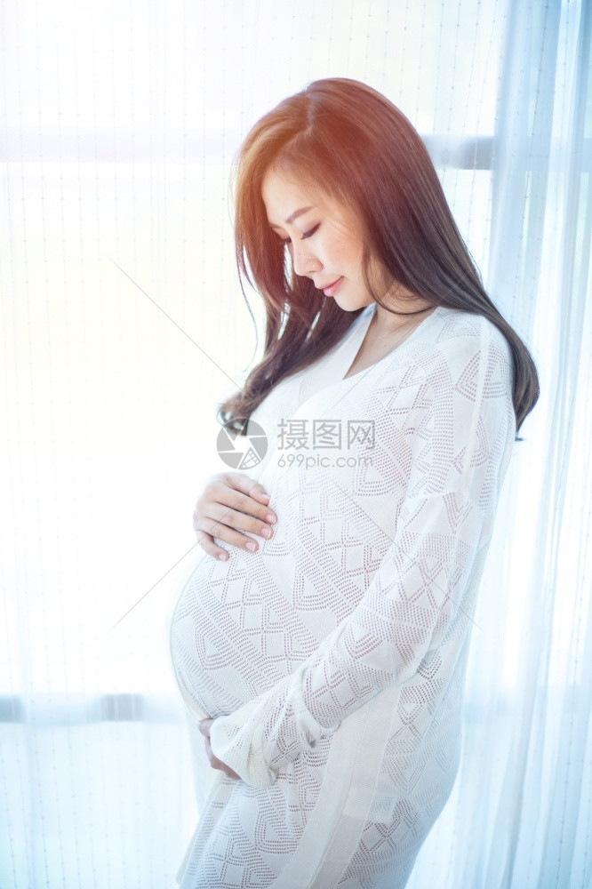 年轻的期待站在家窗户旁边的亚洲年轻美丽孕妇青年在家中的窗户旁边站着母图片