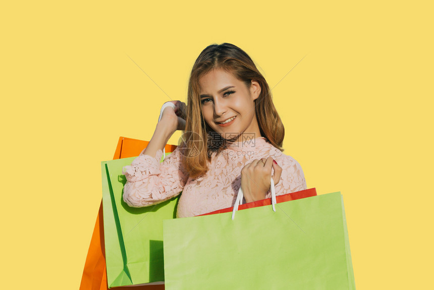 亚裔女美丽孩拿着购物袋在黄色背景上笑着脸在线的卡片移动图片