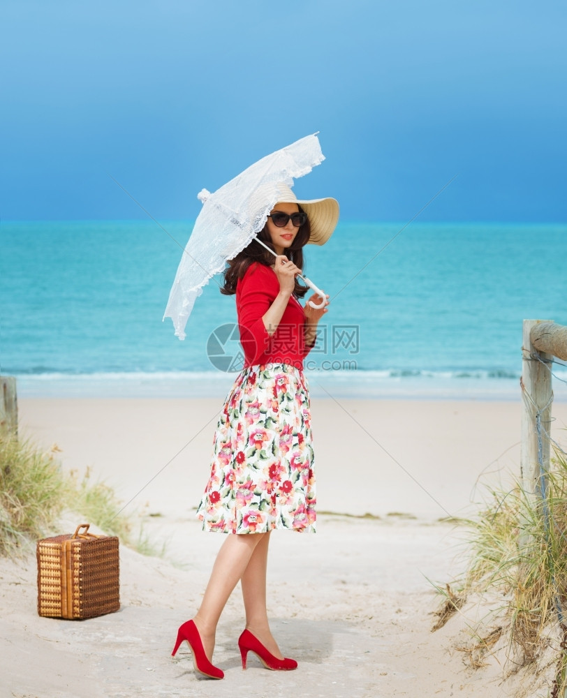 穿红色旅行服的美女在海滩上穿旧式礼服蓝色的复古伞图片