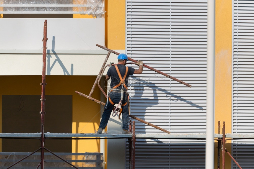 修理家建筑学戴蓝色安全帽的工人在建筑地的脚手架上作一名建筑人在地的脚手架上作图片