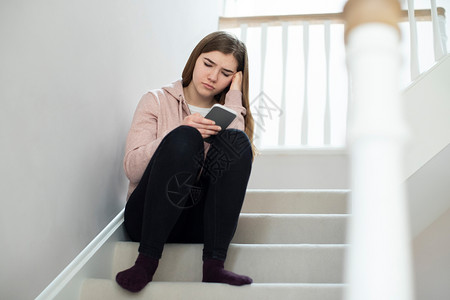 人们网络在家里楼梯上坐着的短信使不快乐的少女受到恐吓存在背景