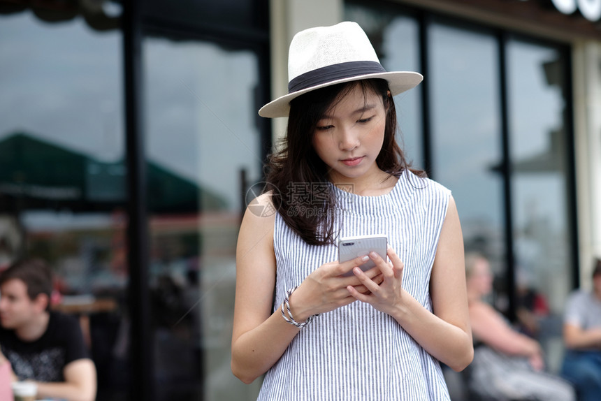 泰国微笑连接使用智能手机在市外咖啡馆前站着户外背景从事技术城市生活方式电话等户外工作的人时使用智能手机的临时风格年轻亚洲女青图片