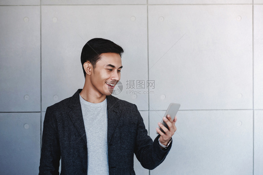 年轻快乐商人的肖像使用智能手机站在工业混凝土墙边通过手机阅读信息和微笑办公室随意的工作图片