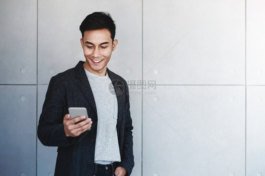 自然互联网专业的年轻快乐商人肖像使用智能手机站在工业混凝土墙边通过手机阅读信息和微笑图片