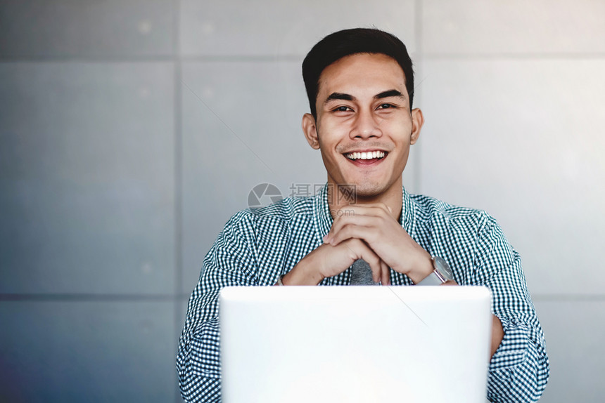 随意的幸福室内快乐亚洲青年商人在工作场所用电脑笔记本工作他的是手放在Chin微笑和看相机图片