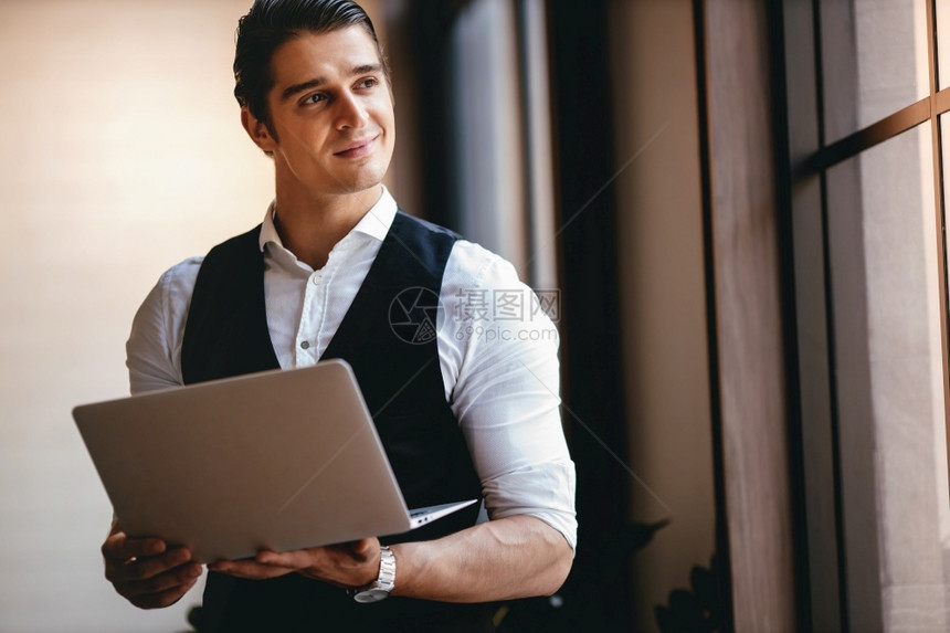 一位年轻的白种商人在现代工作场所使用电脑笔记本微笑的商人站在窗边望着外面成功的首席执行官自信的图片