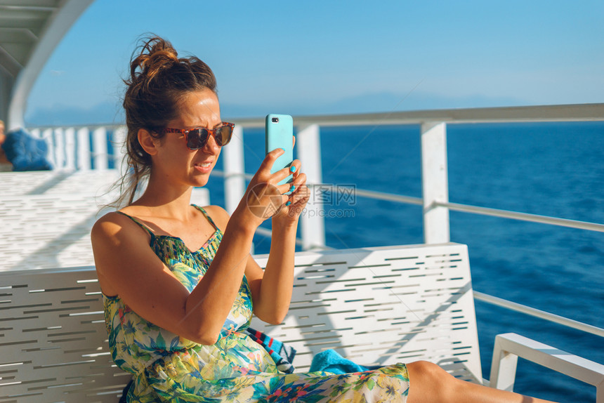 聪明的坐在轮渡船或游艇甲板的长椅上年轻女子在阳光明日度假时使用智能手机照相片拍摄工作环境在假日旅行中用智能手机拍照相片图片