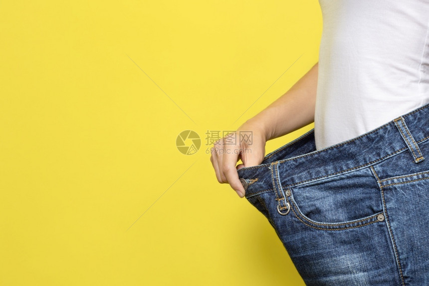 瘦身衣服饮食适当营养减肥的概念显示宽松牛仔裤的苗条女人和她减肥白色T恤和超大号牛仔裤在黄色背景上隔开带有复制空间的横幅饮食减肥的图片