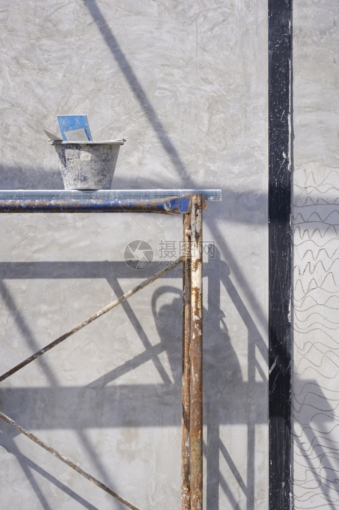 塑料蓝色的水泥桶和石膏工具在垂直框架的房屋建筑工地内水泥墙壁表面架上用阳光和阴影遮的水泥板脚架抹灰图片